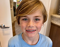 Smiling boy with short, wavy hair wearing a light blue shirt in a bright indoor setting.