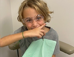 A child with glasses playfully poses with a finger on their lips, sitting in a dental office.