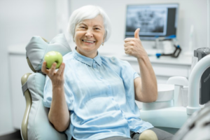 Senior woman sitting in patient chair, holding apple and making thumbs-up